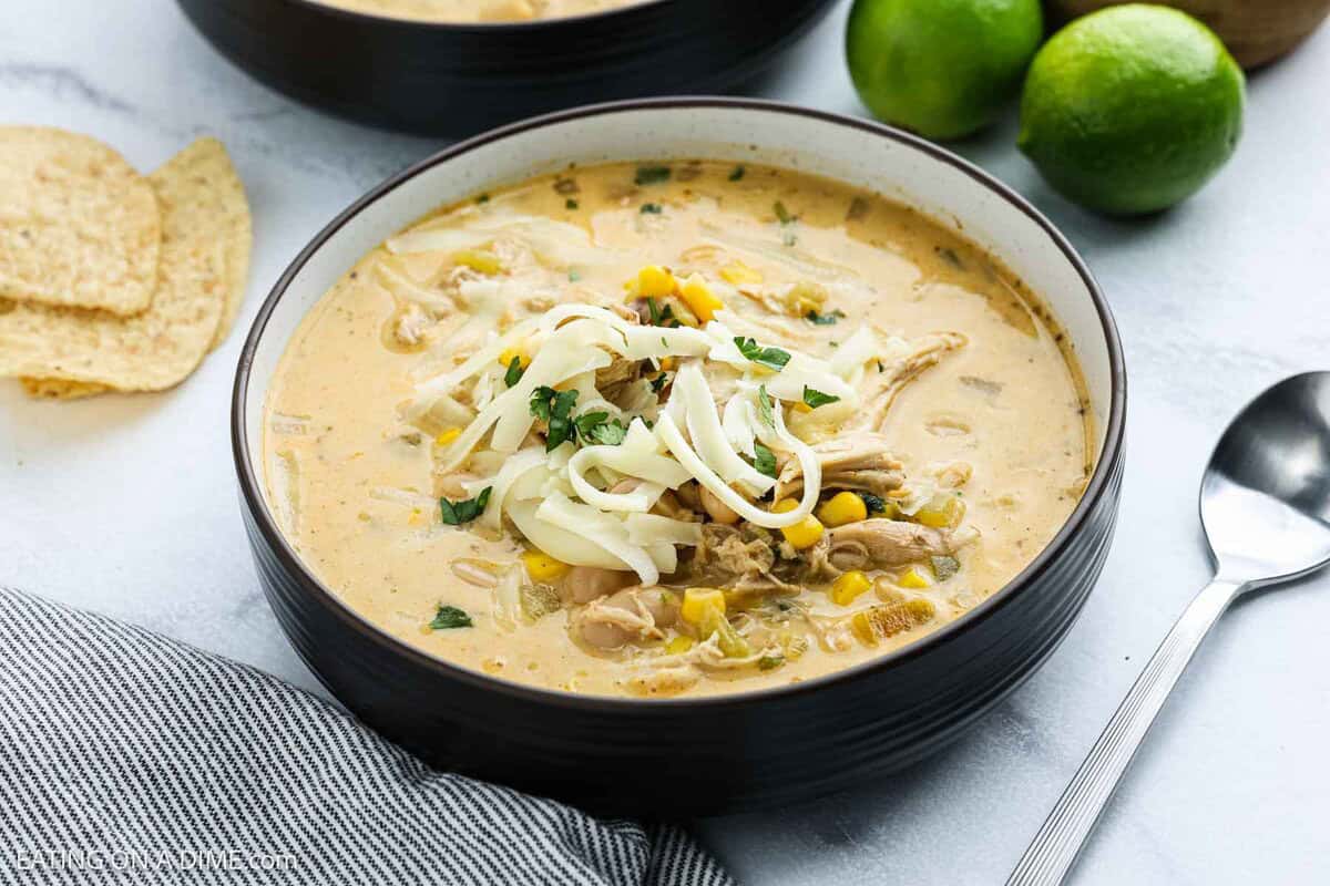 A bowl of creamy White Chicken Chili topped with shredded cheese and herbs, with corn visible in the soup. A spoon, limes, tortilla chips, and a striped napkin are nearby on a white surface.