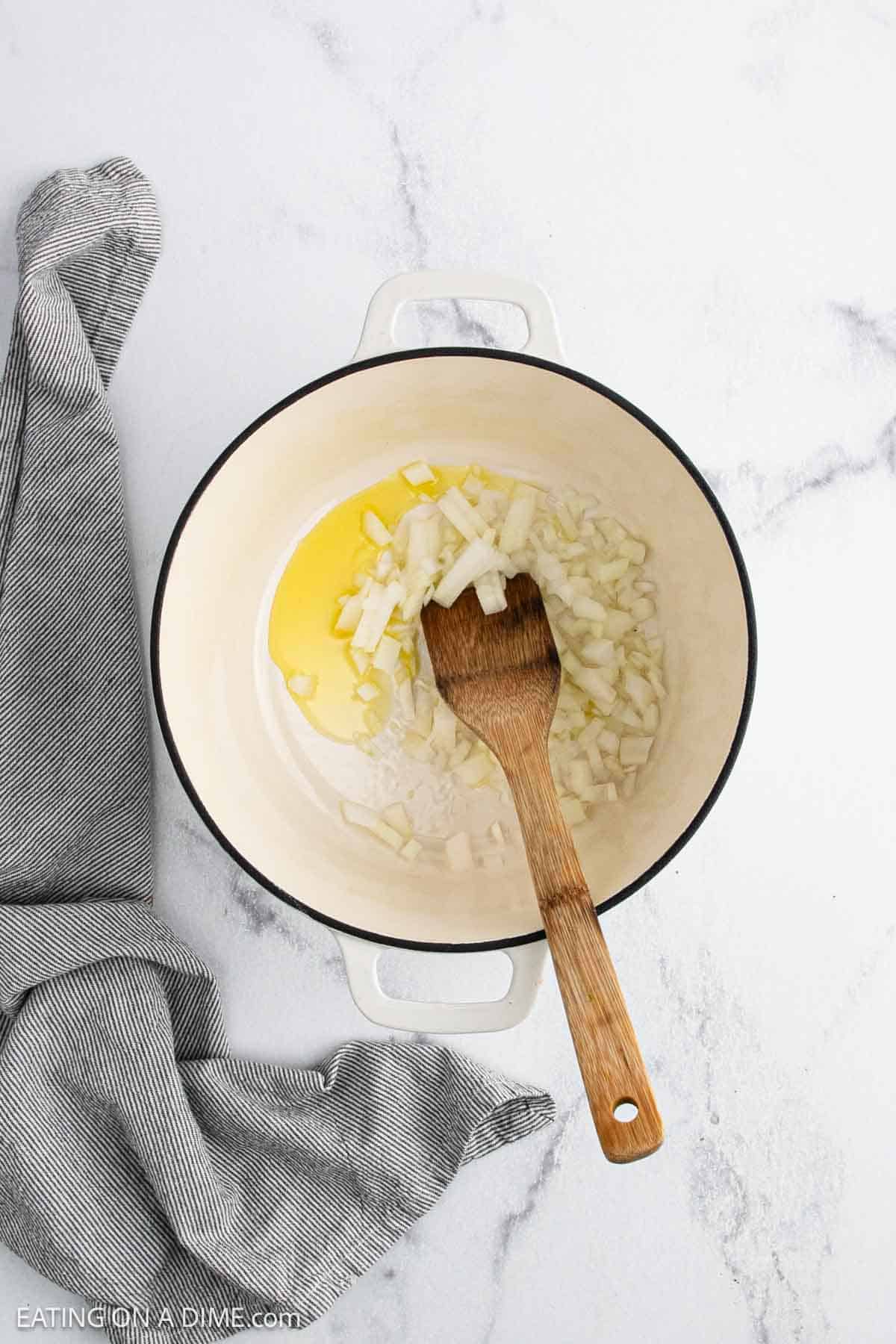 A white pot with chopped onions and oil sits on a white marble surface, ready for White Chicken Chili. A wooden spoon rests in the pot, and a gray-striped cloth is draped nearby.