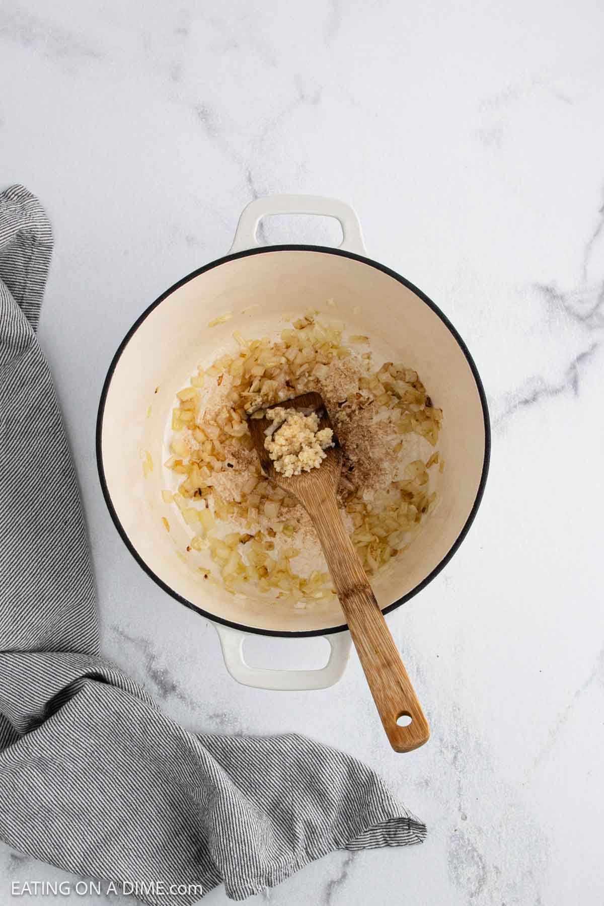 A white Dutch oven sits on a marble surface, filled with sautéed onions and minced garlic—the aromatic base for a flavorful White Chicken Chili—being stirred with a wooden spoon. A gray-striped kitchen towel is nearby.