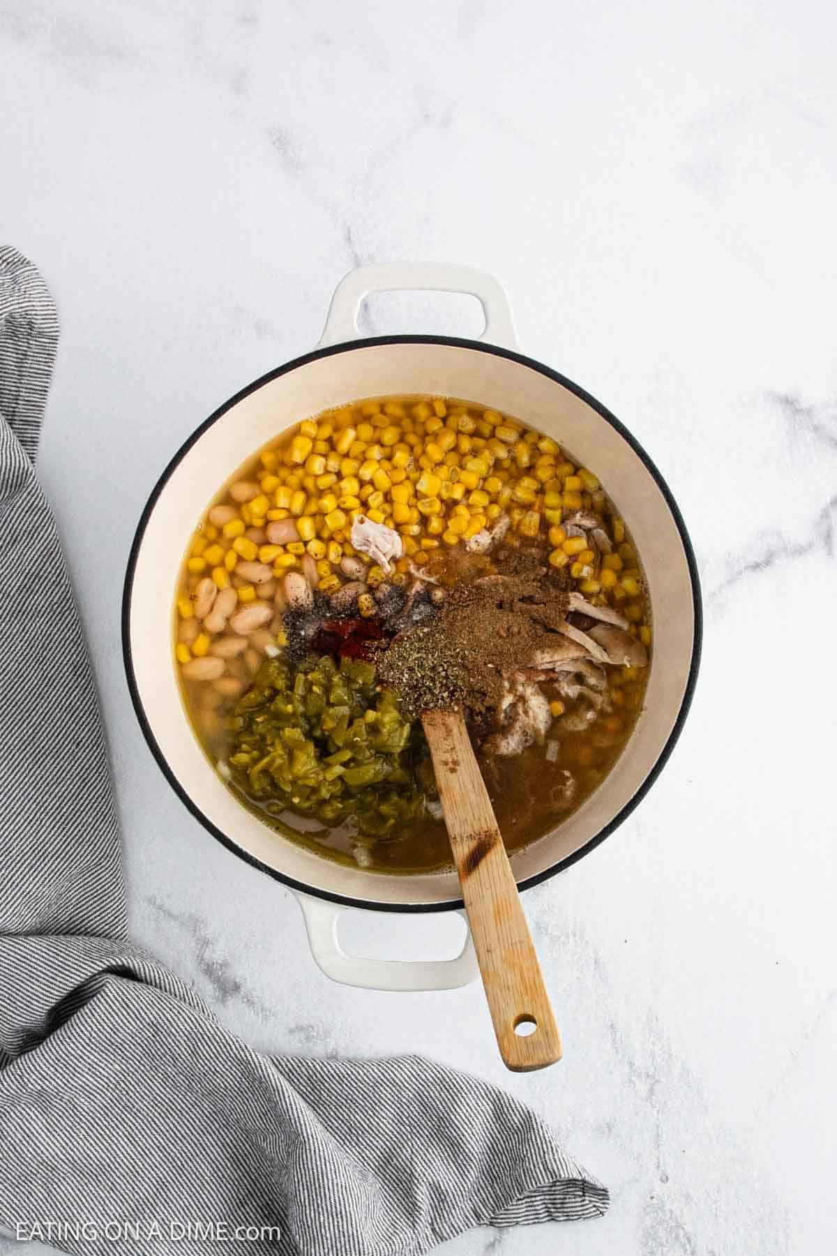 A white pot of White Chicken Chili filled with corn, white beans, green chiles, shredded chicken, seasonings, and broth sits on a white surface with a wooden spoon inside, next to a gray striped cloth.