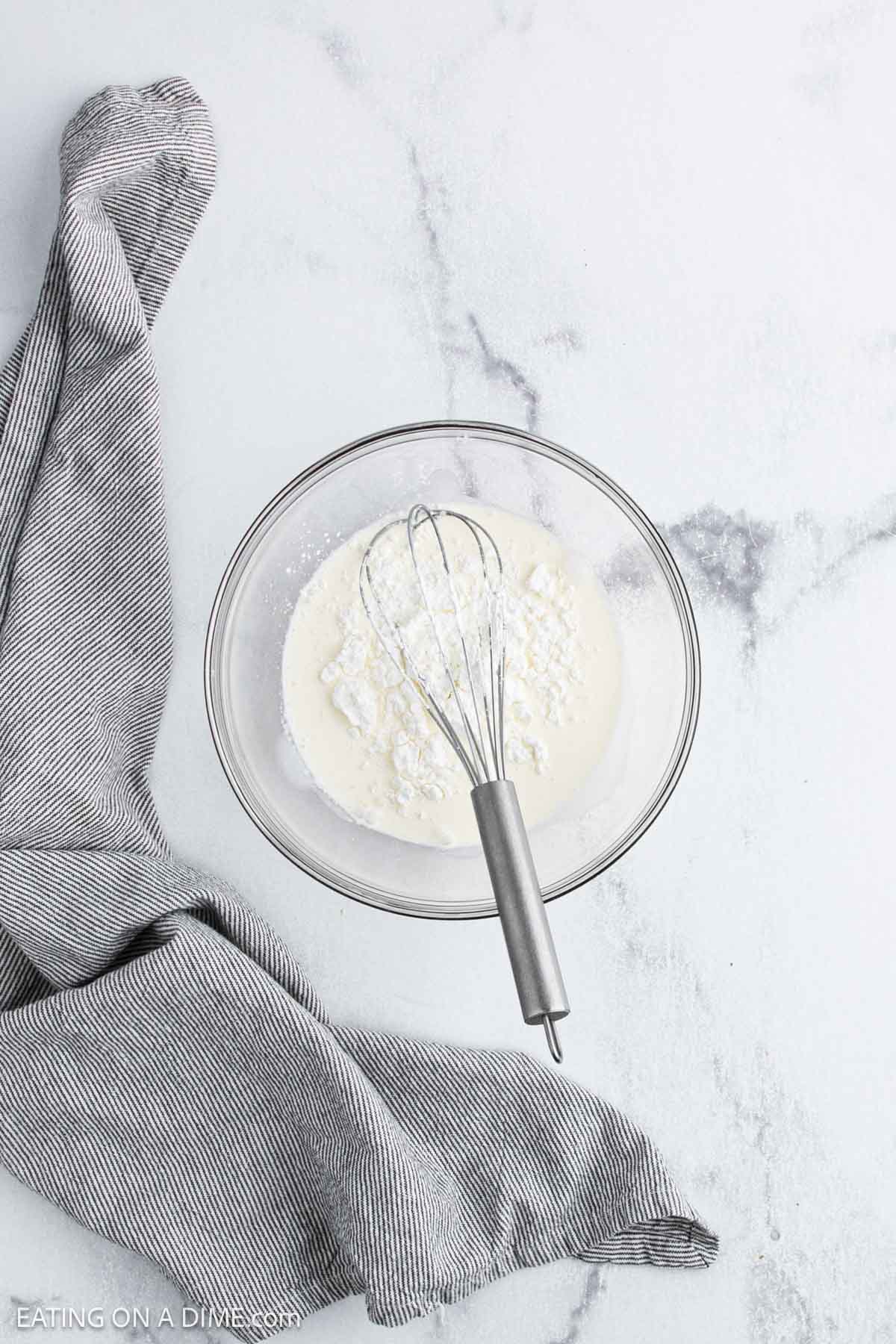 A metal whisk rests in a glass bowl containing a white powdered mixture, perhaps the beginning of a White Chicken Chili recipe. A gray and white striped cloth is draped next to the bowl on a light marble surface.