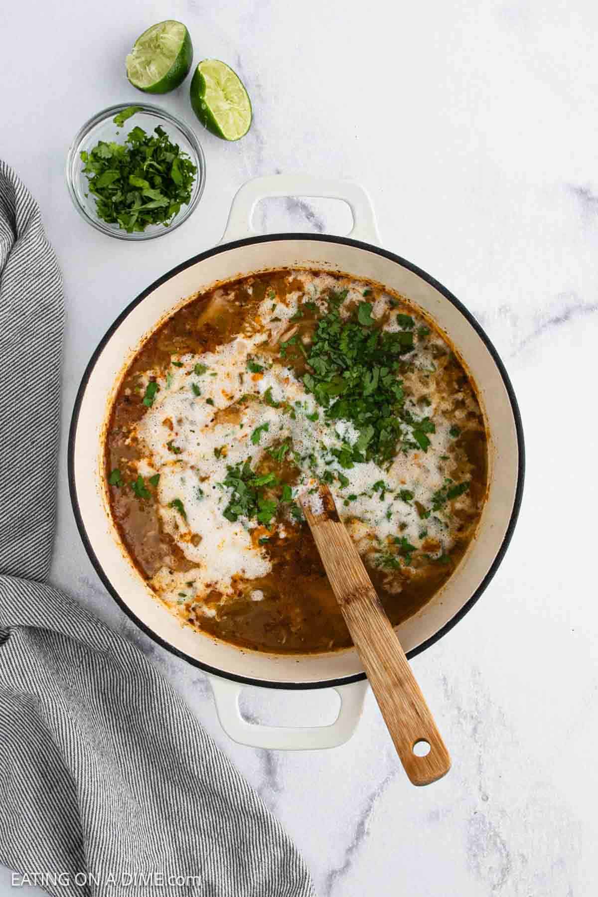 A pot of creamy White Chicken Chili with chopped cilantro on top sits on a marble surface. A wooden spoon rests inside the pot, surrounded by a bowl of cilantro, lime halves, and a gray-striped cloth.