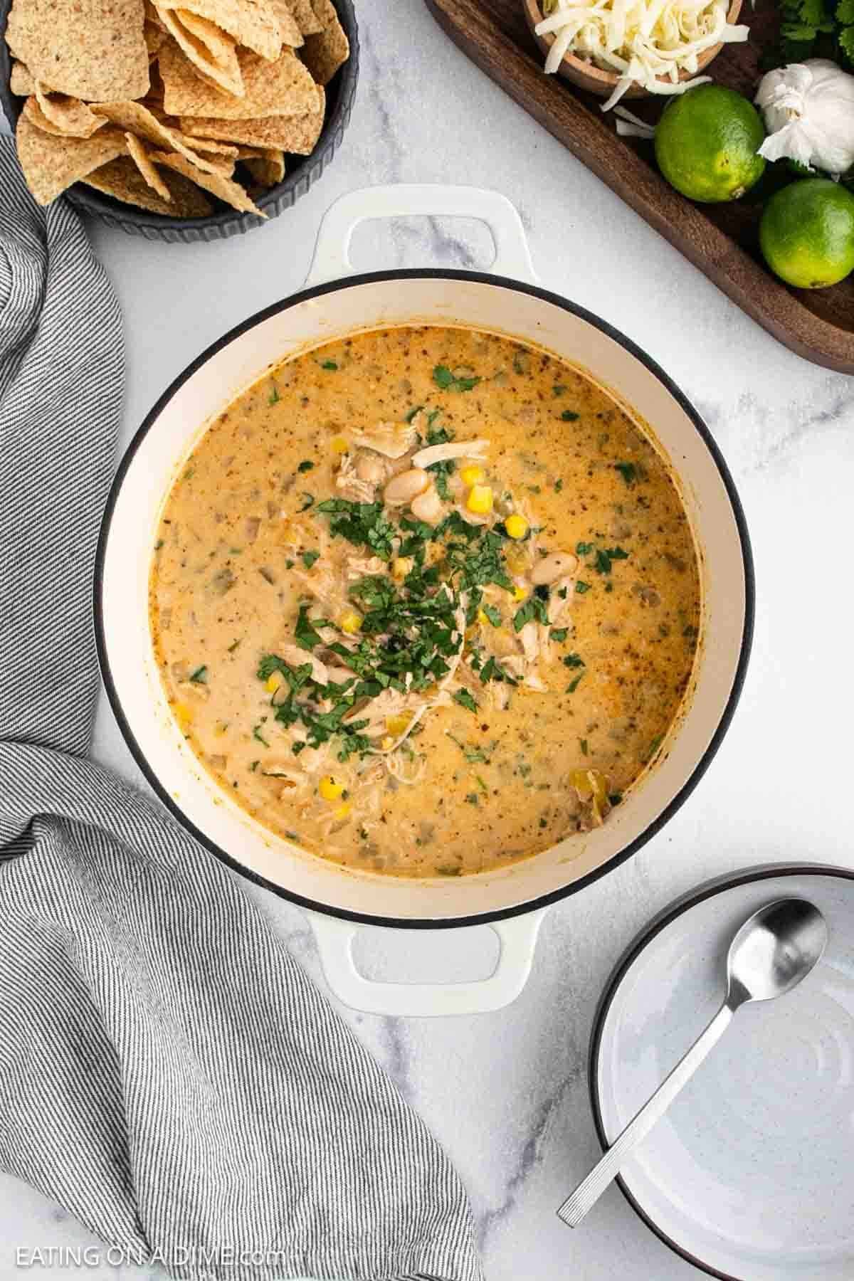 A pot of creamy White Chicken Chili with corn and herbs sits on a white counter, surrounded by tortilla chips, limes, shredded cheese, a striped towel, a spoon, and an empty bowl nearby.