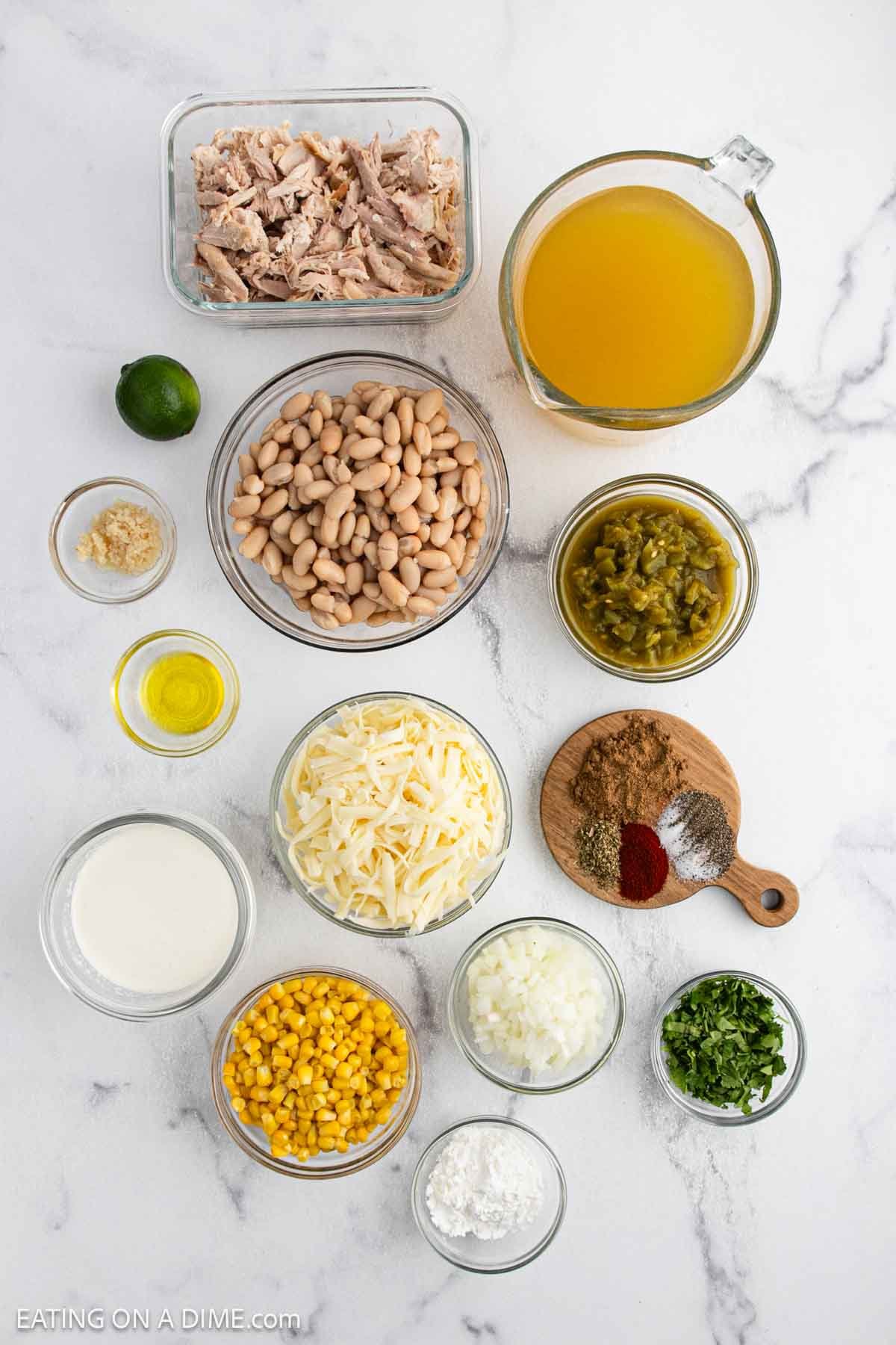 A top-down view of neatly arranged White Chicken Chili ingredients, including shredded chicken, chicken broth, white beans, green chilies, corn, cheese, milk, onion, cilantro, spices, garlic, olive oil, lime, and cornstarch.