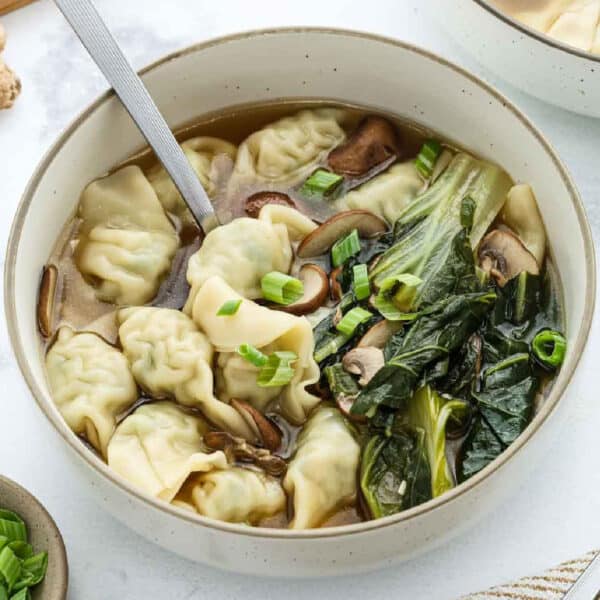 A bowl of Wonton Soup with delicate dumplings, mushrooms, leafy greens, chopped green onions, and a spoon resting inside.