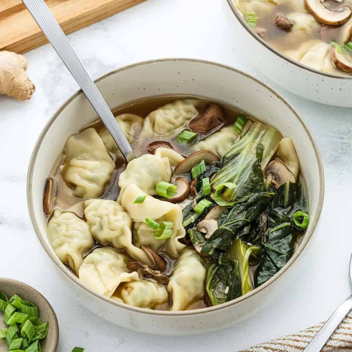 A bowl of wonton soup with dumplings, bok choy, mushrooms, and green onions, with a spoon resting inside. A small bowl of sliced green onions and a piece of ginger are nearby.