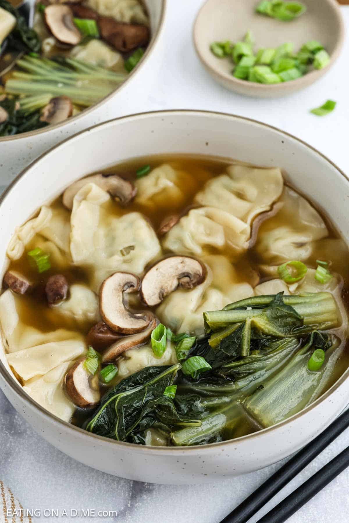 A bowl of wonton soup with dumplings, sliced mushrooms, leafy greens, and chopped green onions in a clear broth, served in a white bowl.