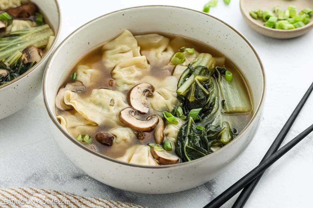 A bowl of wonton soup with dumplings, sliced mushrooms, bok choy, green onions, and broth. Black chopsticks rest nearby and a small bowl of chopped green onions is in the background.