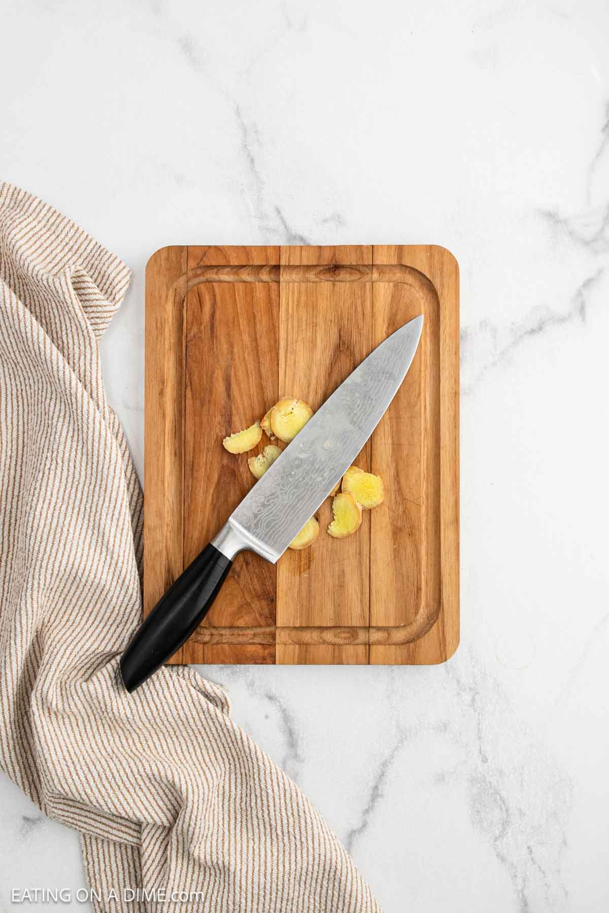 A chef’s knife with a black handle rests on a wooden cutting board with sliced ginger pieces, next to a beige striped kitchen towel on a white marble surface.