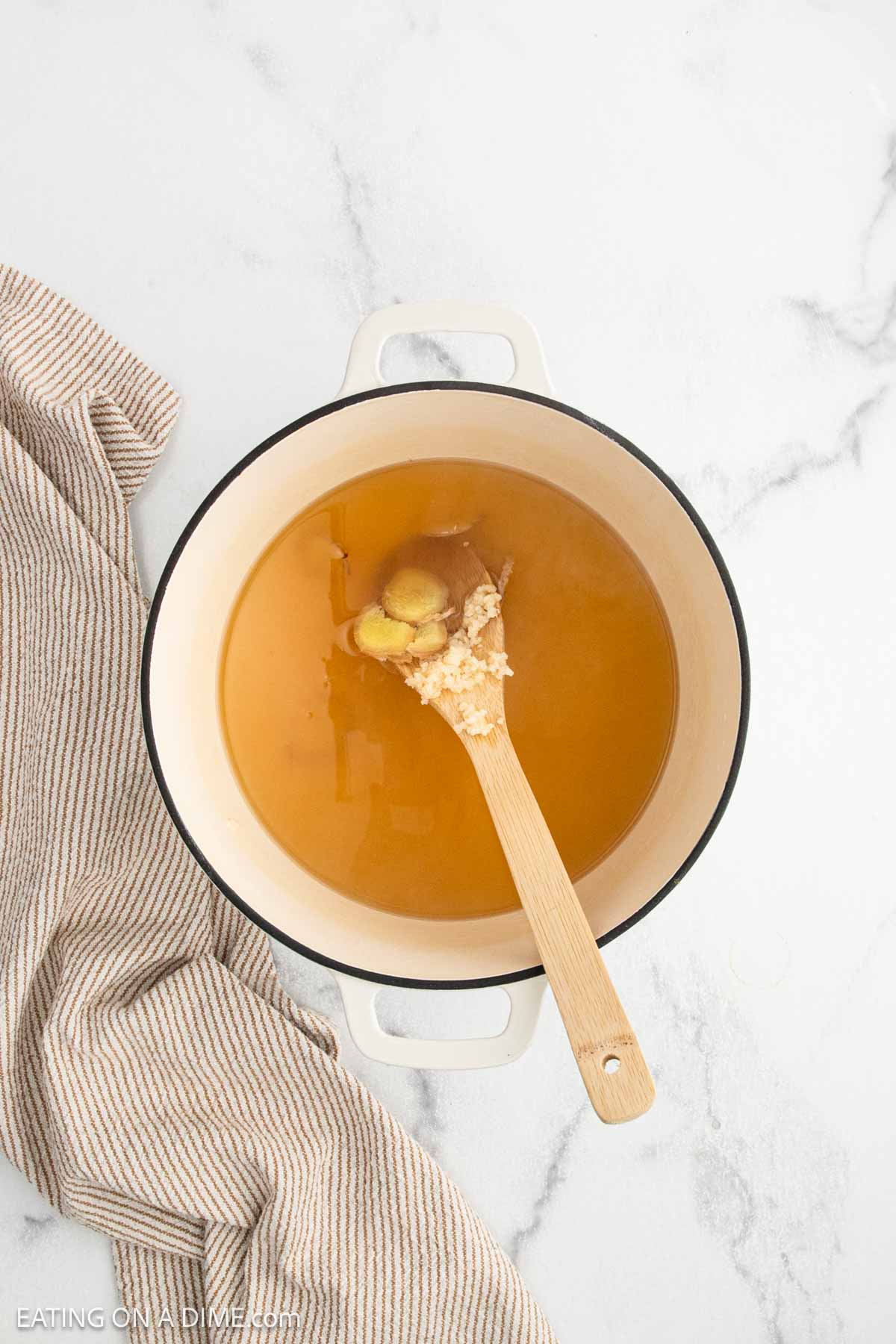 A cream-colored pot filled with light brown broth and a wooden spoon holding ginger and minced garlic, placed on a white marble surface next to a striped kitchen towel.