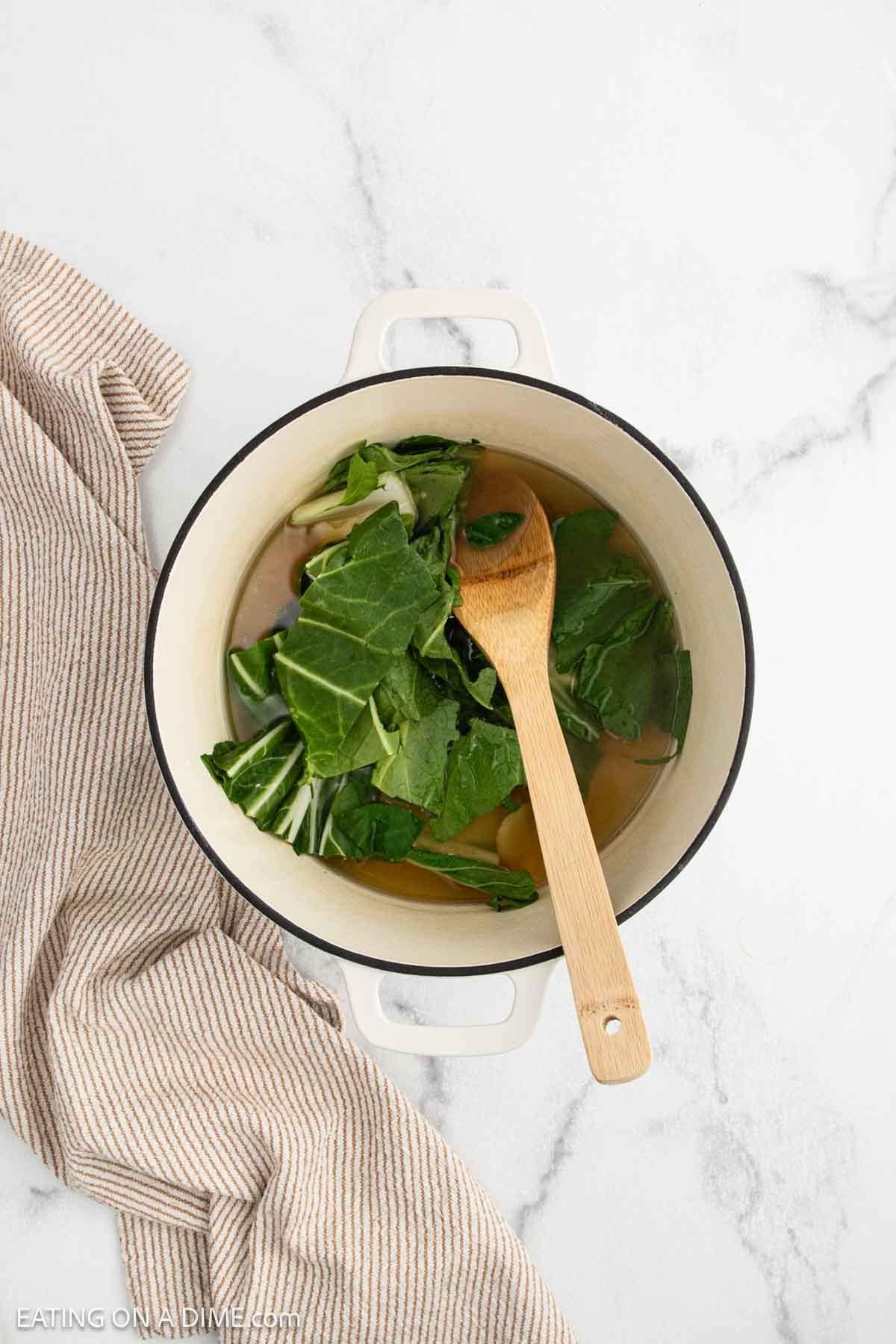 A white pot filled with leafy greens and broth sits on a marble surface, accompanied by a wooden spoon and a beige striped towel.