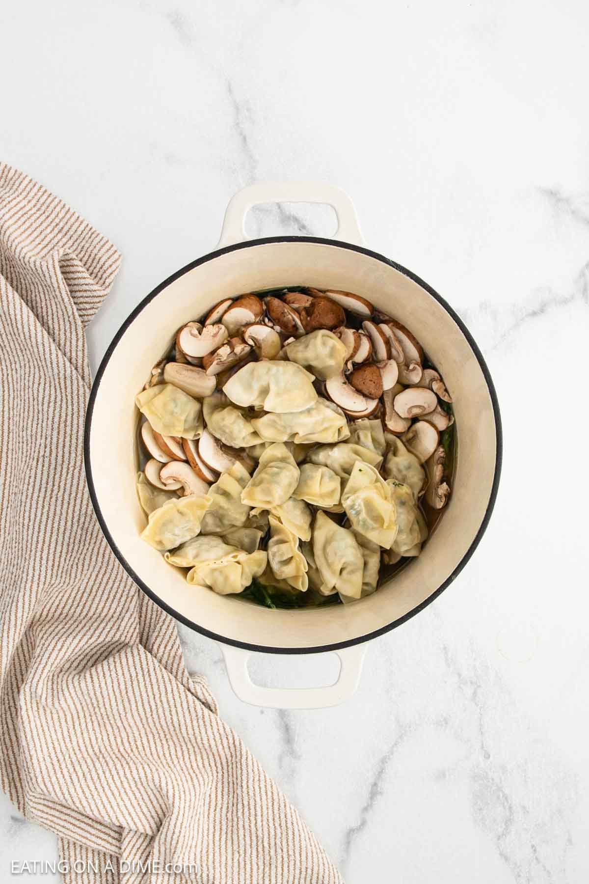 A white pot filled with uncooked dumplings and sliced mushrooms sits on a marble surface next to a beige and white striped towel.