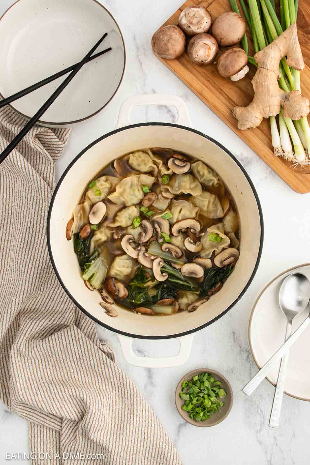 Top-down view of a pot of dumpling soup with mushrooms, greens, and broth. Nearby are a striped towel, serving bowls with chopsticks and spoons, and a cutting board with mushrooms, green onions, and ginger.