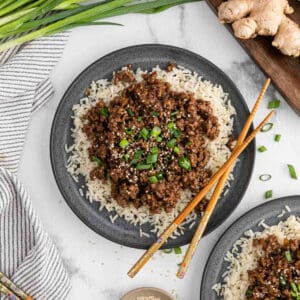 A plate of white rice topped with ground beef, green onions, and sesame seeds forms a classic Korean Beef Bowl. Chopsticks rest on the plate, with fresh ginger, green onions, and a striped napkin nearby on the marble surface.
