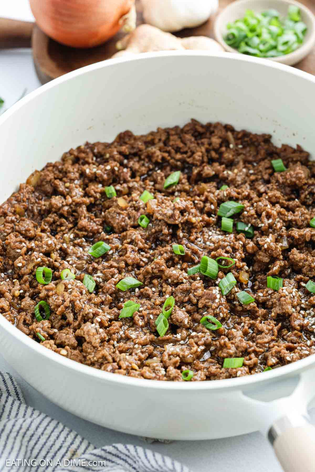 Cooked ground beef for Korean Beef Bowls is topped with sliced green onions and sesame seeds in a white skillet, with an onion, mushrooms, garlic, and chopped green onions in the background.