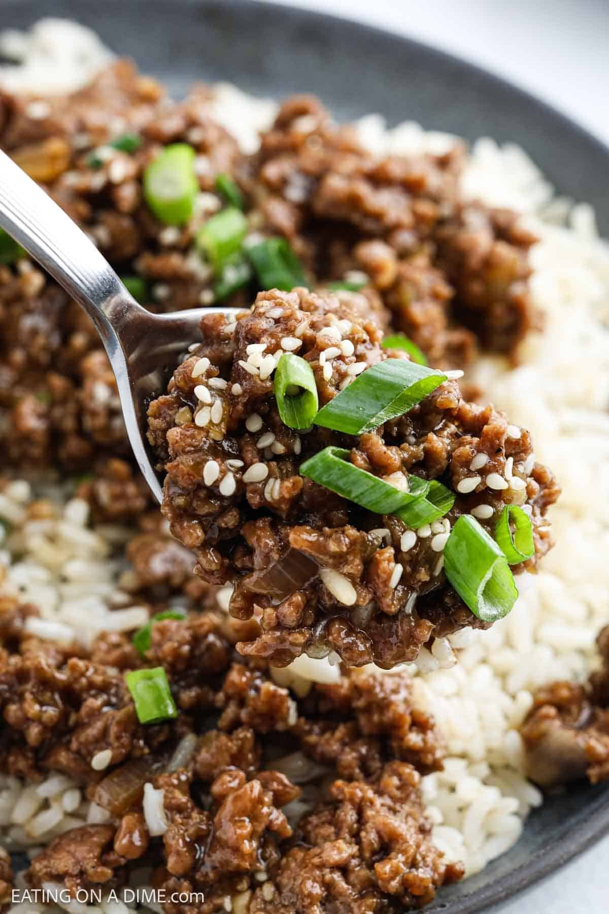 A close-up of a fork holding a scoop of Korean Beef in a savory sauce, topped with sesame seeds and chopped green onions, served over a bed of white rice.
