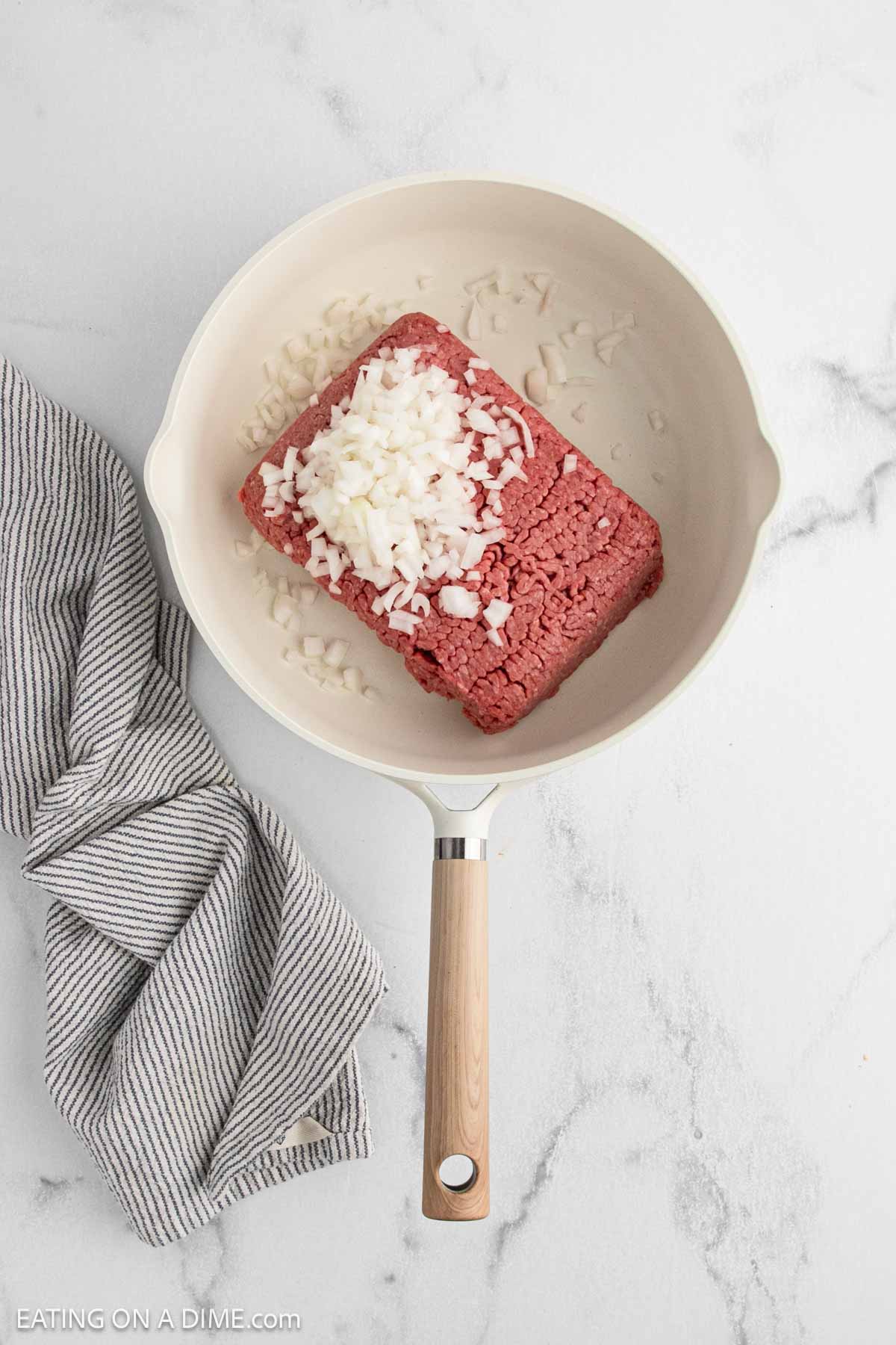 A white skillet with raw ground beef and chopped onions sits on a marble countertop next to a folded gray and white striped kitchen towel, ready to be transformed into a delicious Korean Beef Bowls recipe.
