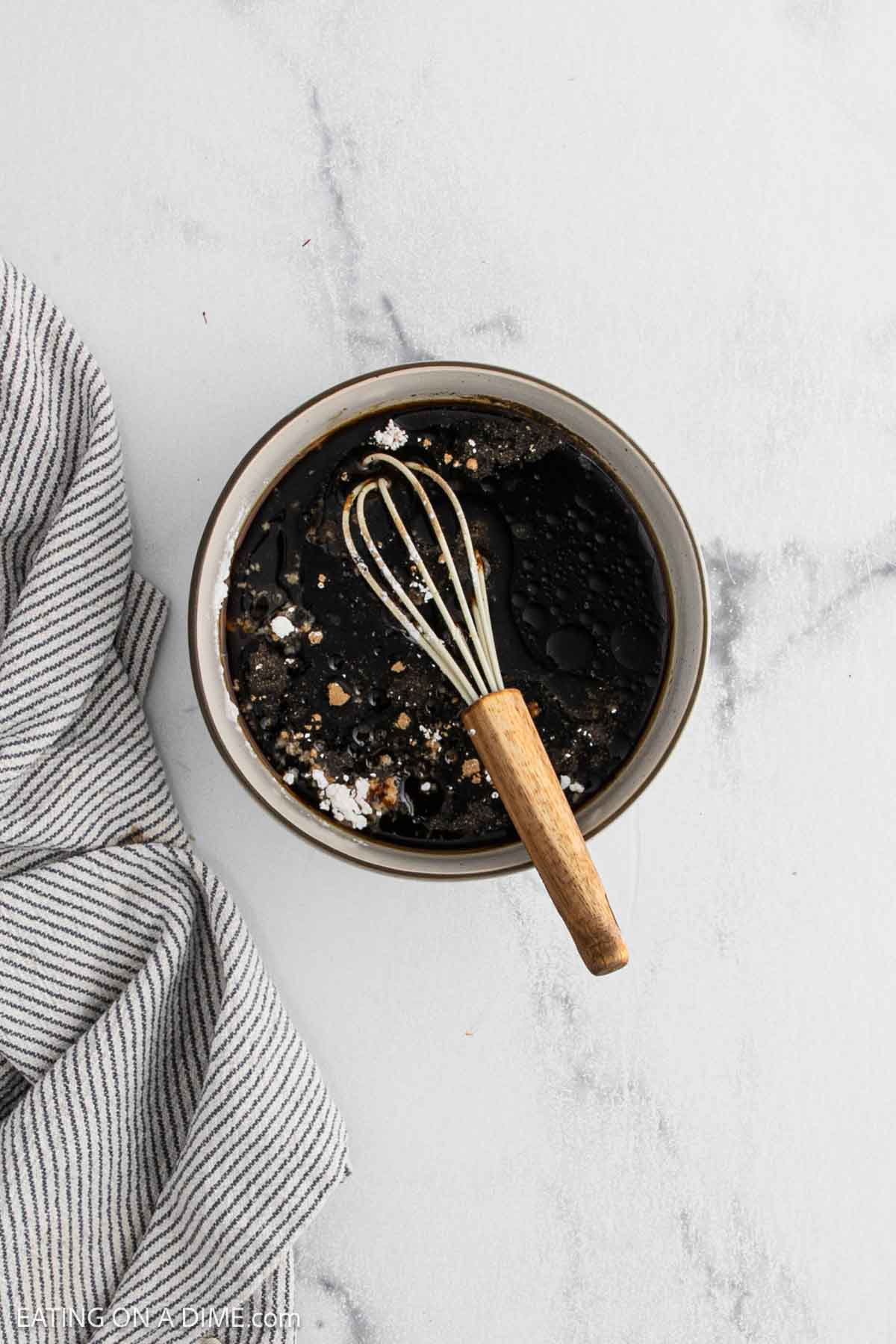 A bowl filled with a dark liquid mixture and seasonings, perfect for a Korean Beef Bowls recipe, with a small whisk resting inside. A gray and white striped cloth is placed next to the bowl on a light marble surface.