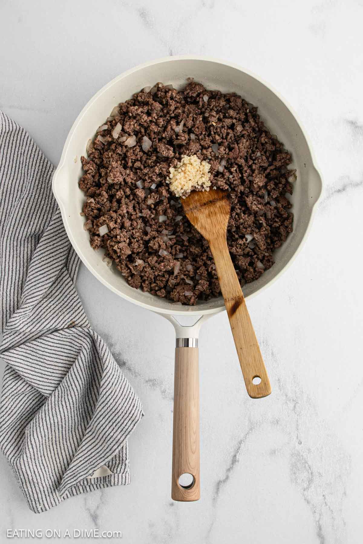 A skillet with cooked ground beef and chopped onions, perfect for Beef Bowls, is topped with minced garlic. A wooden spatula rests in the pan, and a striped kitchen towel lies beside it on a white marble surface.