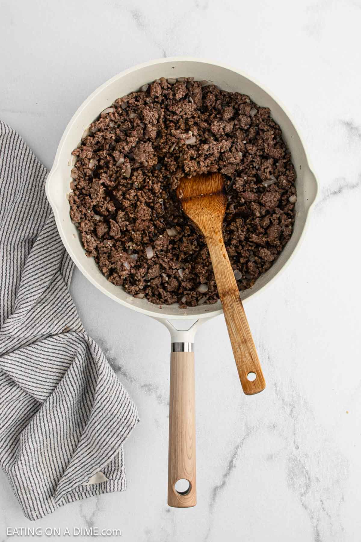 A white skillet filled with cooked ground beef and diced onions sits on a marble surface, perfect for a delicious Korean Beef Bowls recipe. A wooden spoon rests in the skillet, and a striped dish towel is nearby.
