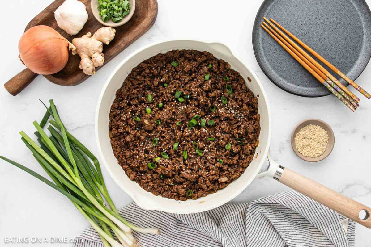 A skillet filled with cooked ground beef garnished with chopped green onions, surrounded by fresh green onions, an onion, ginger, sesame seeds, and chopsticksโa delicious Korean Beef Bowls recipe ready to enjoy on a white surface.