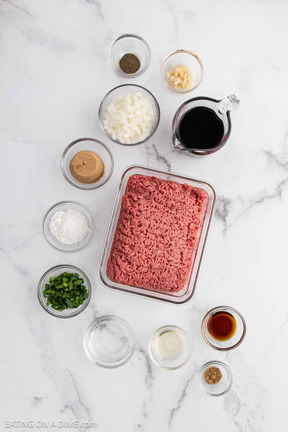 A tray of raw ground beef is surrounded by small bowls of chopped onions, minced garlic, and classic ingredients for an easy Korean Beef Bowls recipe on a white surface.