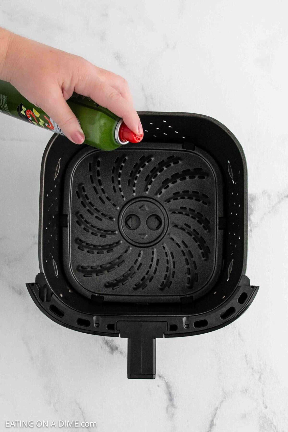 A hand sprays oil from a green can into the basket of a black air fryer on a white marble surface, prepping it to cook crispy gnocchi.
