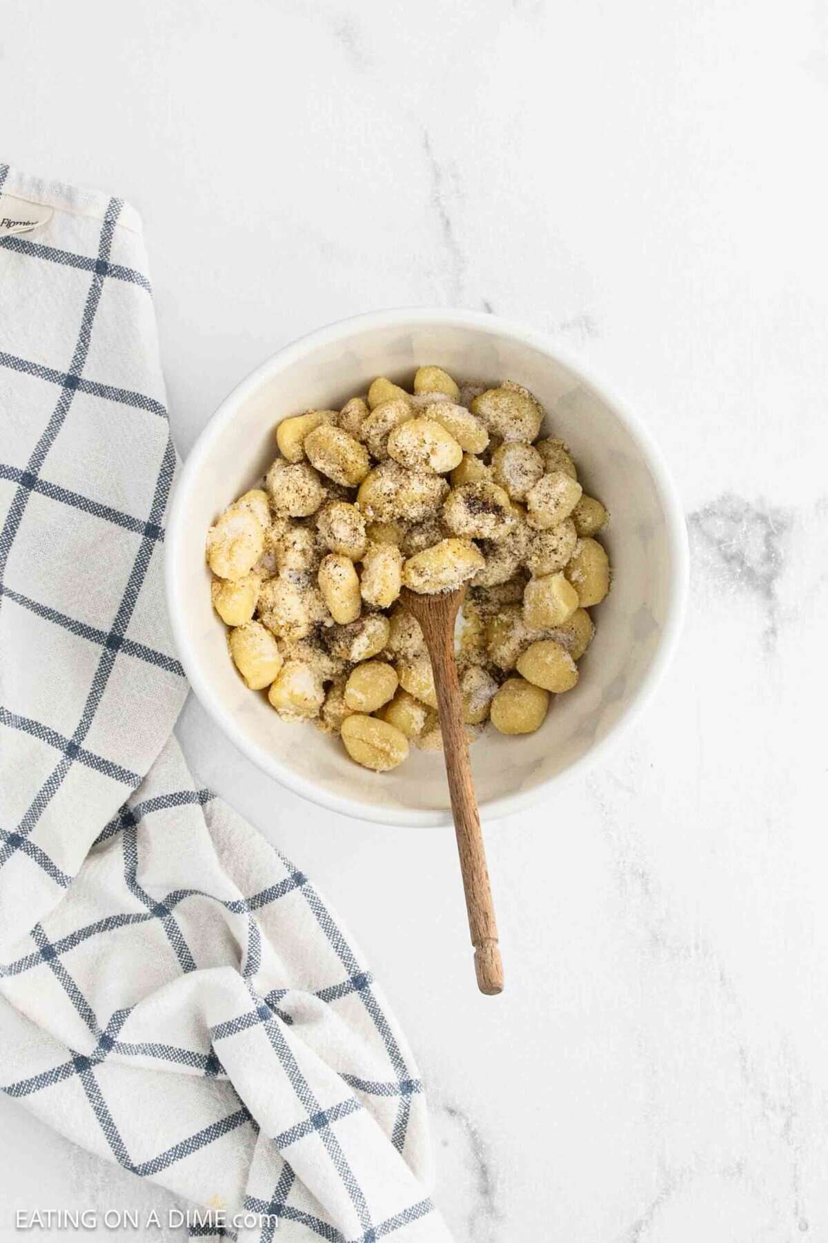 A white bowl filled with Air Fryer Gnocchi, topped with grated cheese and pepper, with a wooden spoon resting inside. The bowl sits on a white surface next to a blue and white checkered cloth.