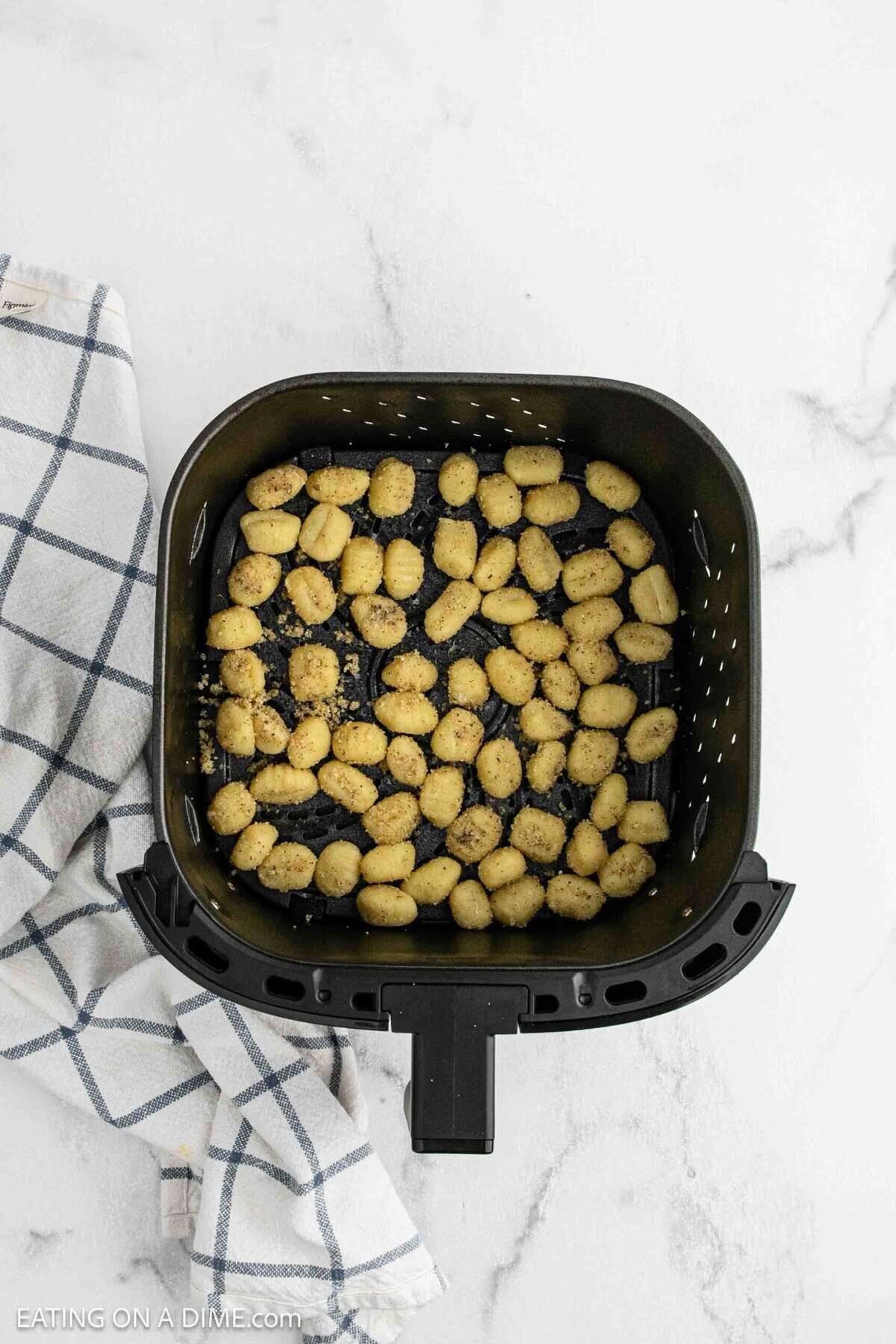 A black air fryer basket filled with seasoned gnocchi sits on a white marble surface next to a folded white and blue checkered kitchen towel, making it a delicious example of an easy Air Fryer Gnocchi recipe.