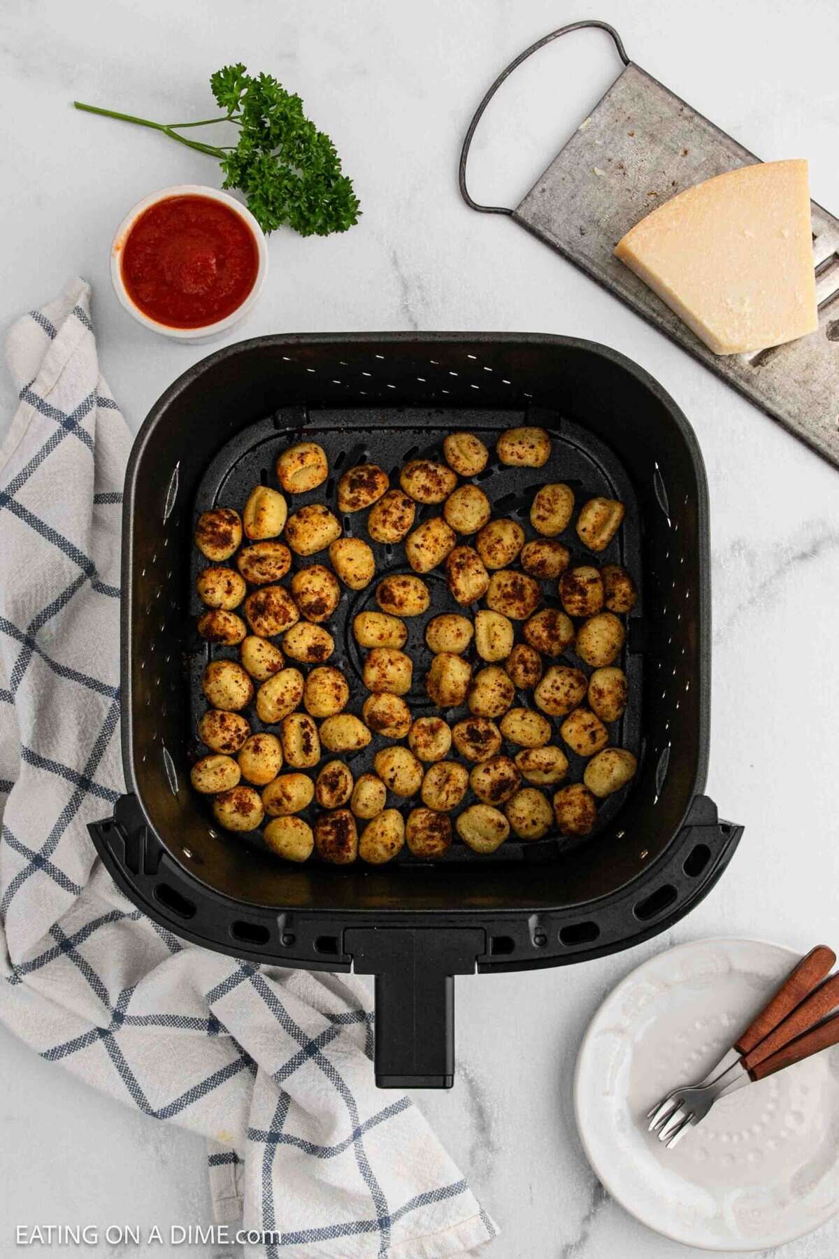 Air fryer basket filled with golden brown Air Fryer Gnocchi on a marble countertop. A bowl of marinara sauce, Parmesan wedge, parsley, towel, and empty white plates with forks are arranged nearby for the perfect gnocchi recipe setup.