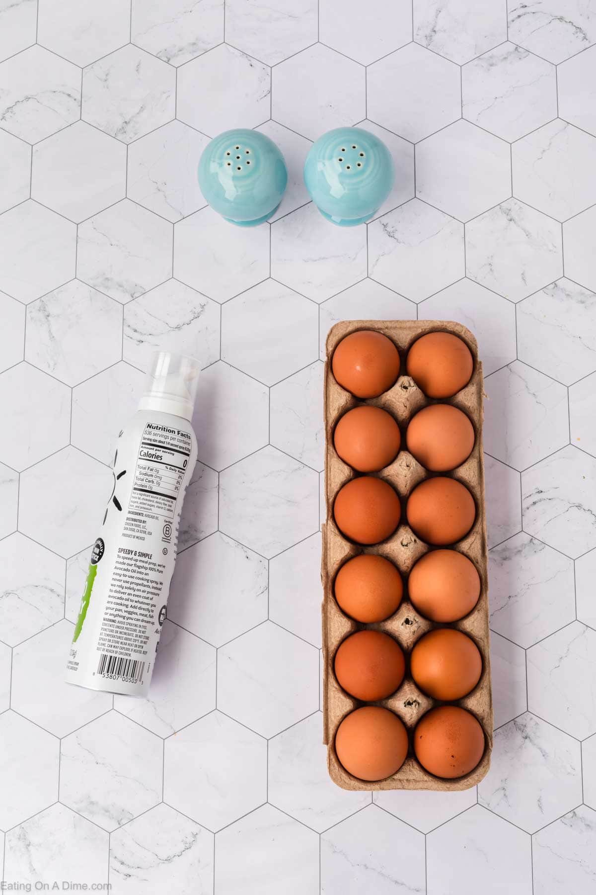 A carton of ten brown eggs, a can of cooking spray, and two blue salt and pepper shakers are arranged on a white hexagon-tiled surface.