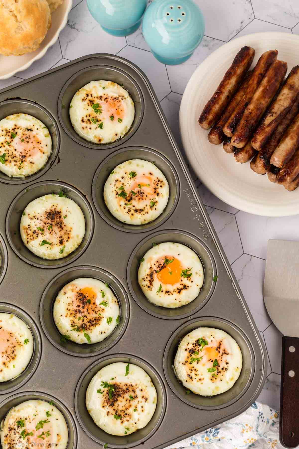 A muffin tin filled with baked eggs garnished with herbs sits on a table next to a plate of cooked sausages, salt and pepper shakers, a biscuit, and a metal spatula.