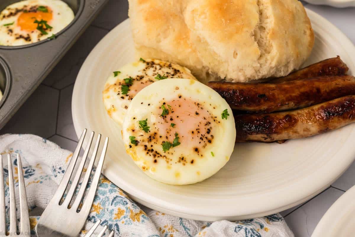 A plate with two baked egg rounds sprinkled with herbs, two sausage links, and a biscuit. A fork and napkin are nearby, and a muffin tin with baked eggs is partially visible in the background.