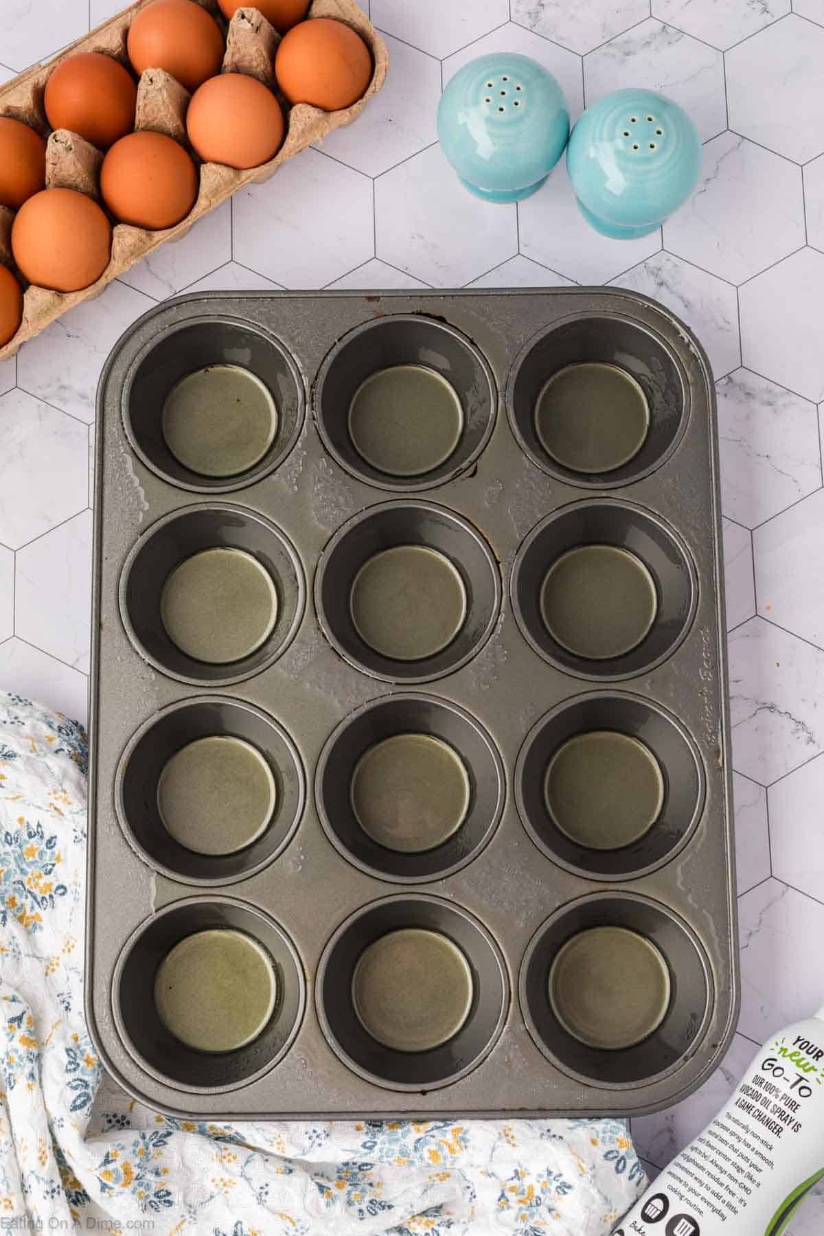 A 12-cup muffin tin sprayed with oil sits on a counter next to a carton of brown eggs, two blue salt and pepper shakers, and a floral cloth on a white hexagon-patterned surface.