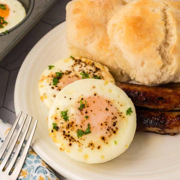 A plate with two baked eggs topped with pepper and herbs, two pieces of sausage, and two fluffy biscuits. A fork and a patterned napkin are beside the plate.