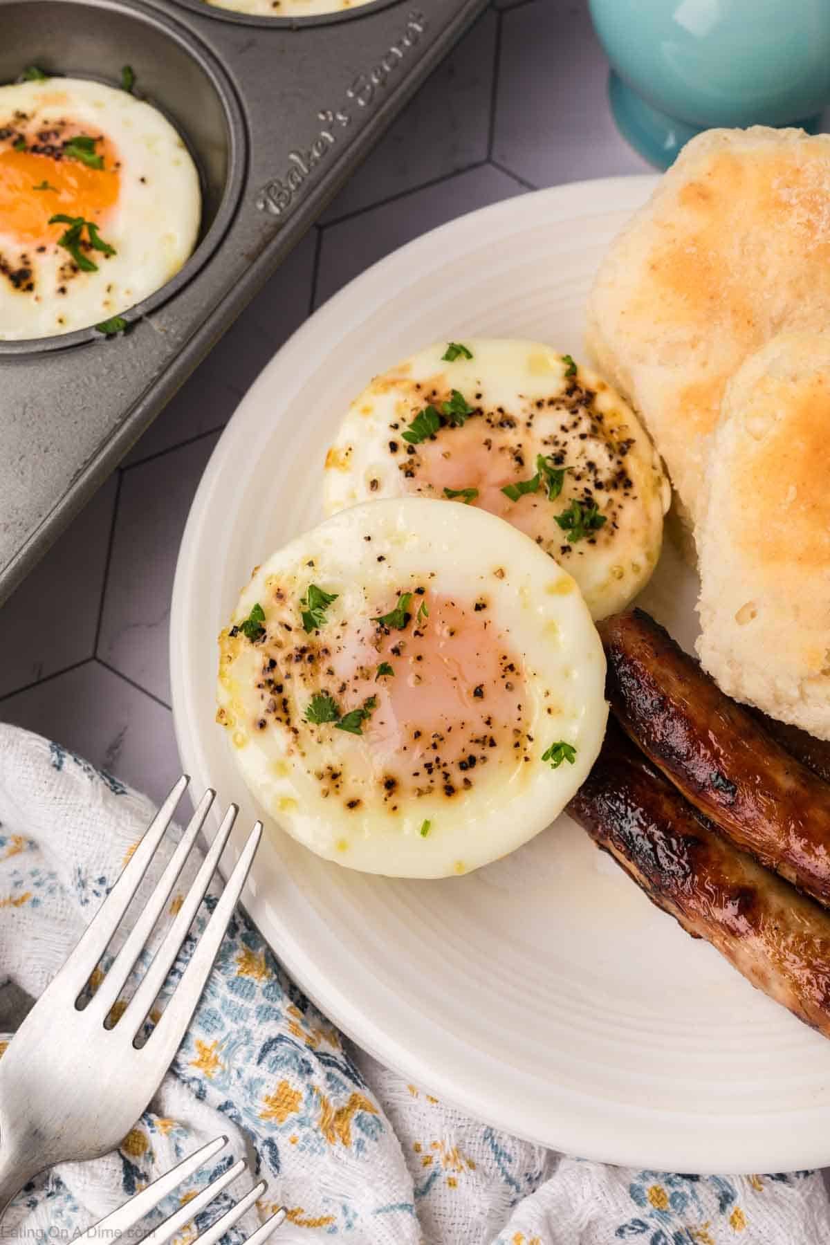 A plate with two baked eggs topped with herbs, two sausage links, and biscuits. A muffin tin with more baked eggs is nearby, and a floral napkin with a fork is in the corner.