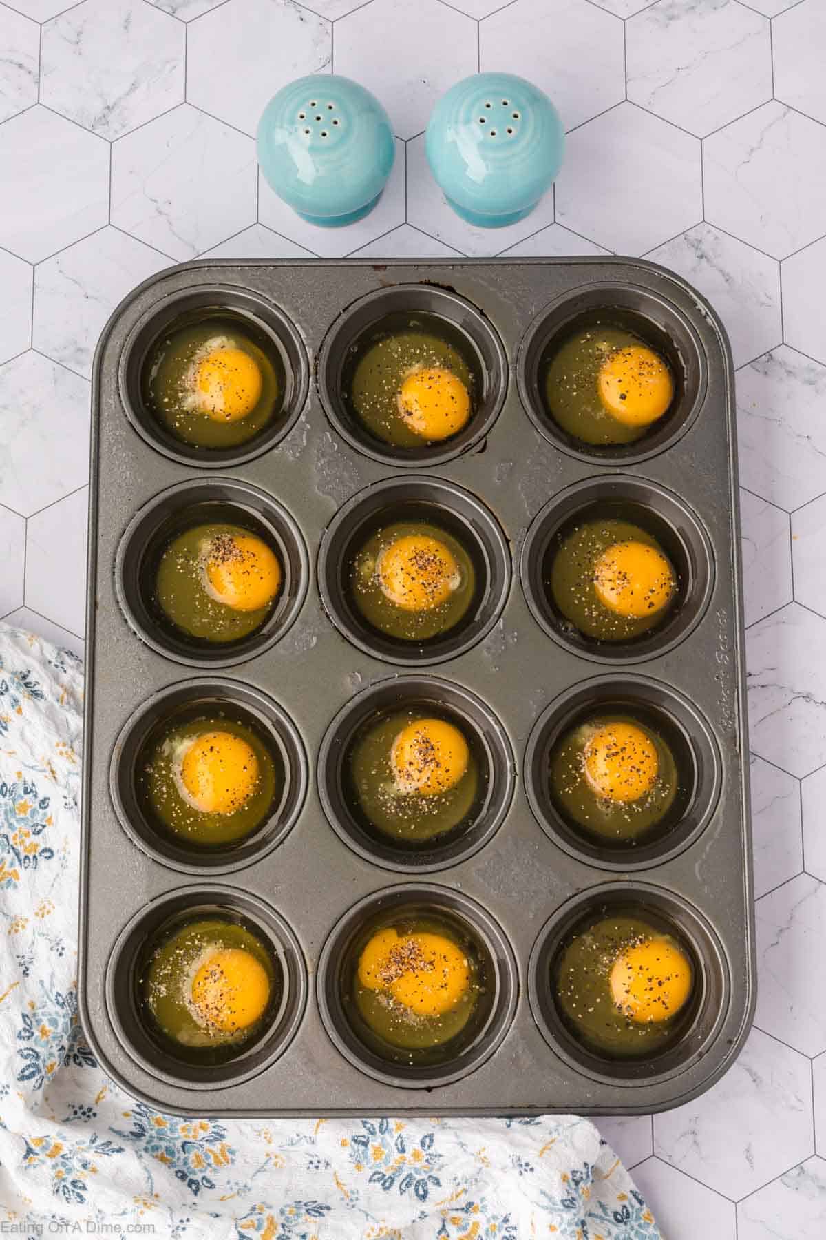 A muffin tin holds twelve raw eggs, each topped with black pepper, on a hexagon-tiled countertop. Two blue salt and pepper shakers and a floral cloth are nearby.
