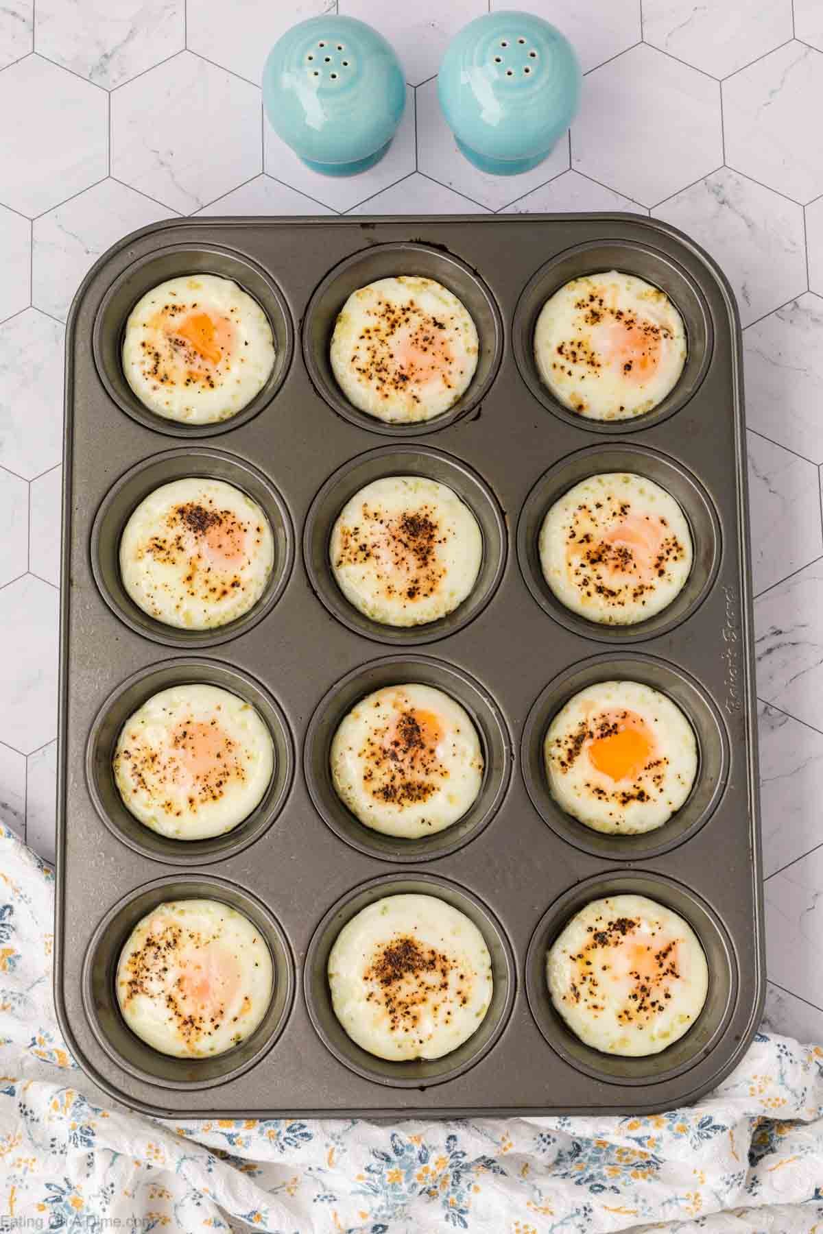 A muffin tin with twelve baked eggs, each topped with seasoning, sits on a light patterned surface. Nearby are two blue salt and pepper shakers and part of a floral cloth.