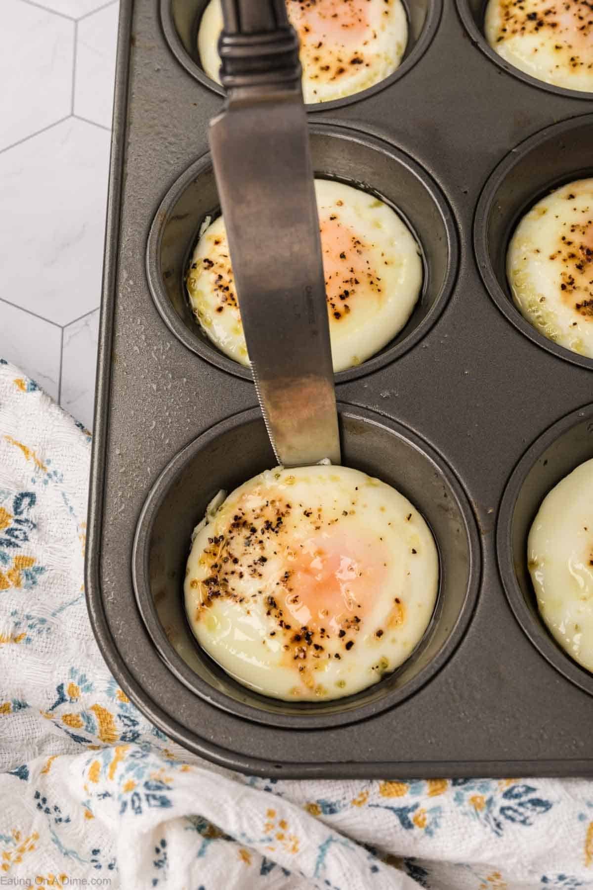 A metal knife loosens a baked egg from a muffin tin, with several other baked eggs in the pan. A floral-patterned towel is nearby on the counter.