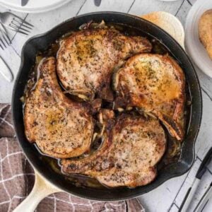 A cast iron skillet filled with four seasoned, cooked pork chops in a savory sauce, featured as part of Meal Plan 83, set on a rustic wooden table with a brown kitchen towel and dining utensils nearby.