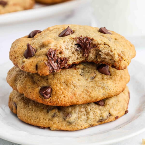 Three chocolate chip cookies are stacked on a white plate. The top cookie, inspired by a banana cookie recipe, has a bite taken out, revealing melted chocolate chips inside. The cookies look soft and chewy.