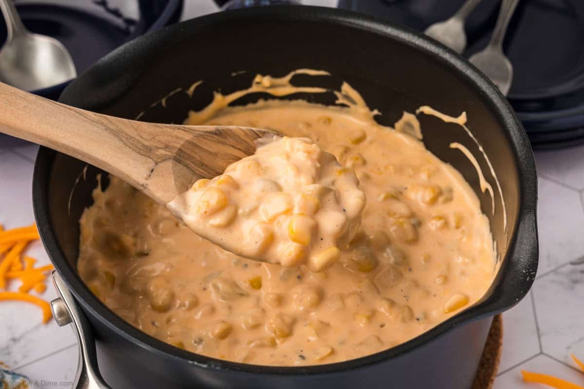 A wooden spoon lifts a serving of creamy, cheesy corn from a black saucepan filled with this irresistible Cheesy Corn Recipe. Shredded cheese and blue plates with spoons are visible in the background.