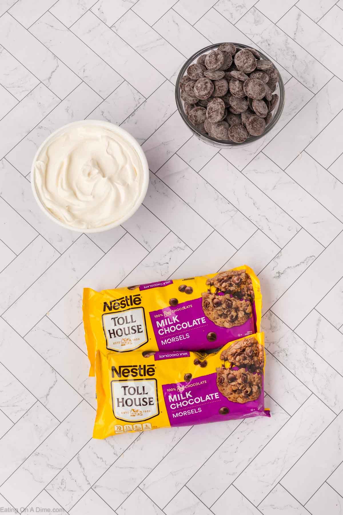Two bags of Nestlรฉ Toll House milk chocolate morsels, a glass bowl of chocolate chips, and a bowl of Cool Whip are arranged on a white herringbone-patterned surface.