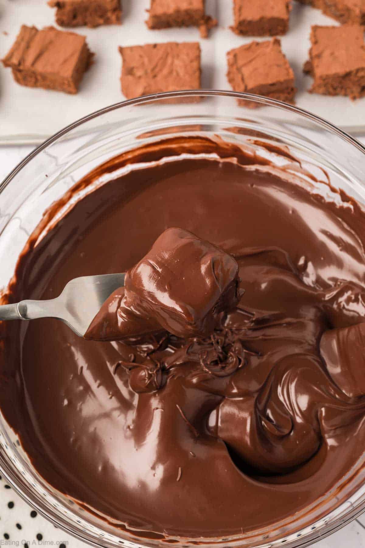 A fork holds a brownie piece dipped in smooth, melted chocolate, turning this dessert into a decadent candy treat, with more chocolate coating in a glass bowl below and brownie squares on a tray in the background.