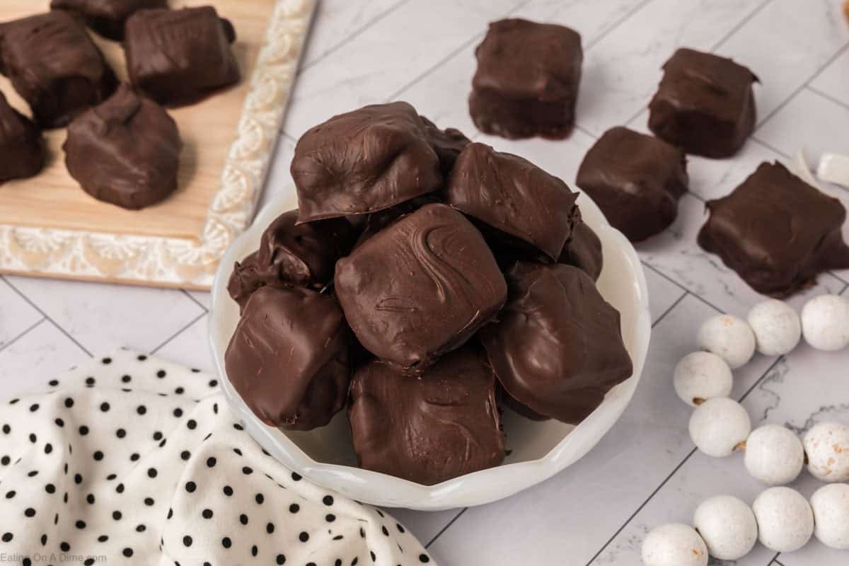 A white bowl filled with chocolate-covered Cool Whip candy squares sits on a marble surface, with more candy pieces scattered around and on a wooden tray nearby. A white polka dot cloth and some white candies are also visible.