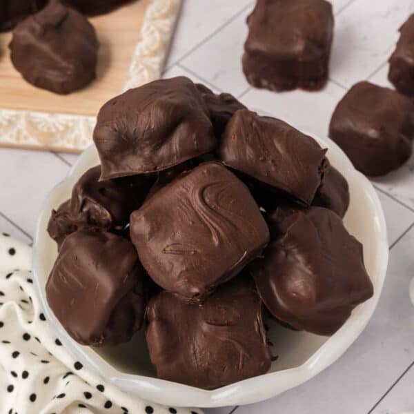 A white bowl filled with homemade Cool Whip candy squares sits on a white countertop, with more candy scattered nearby and on a wooden board in the background. A white cloth with black dots is partially visible.