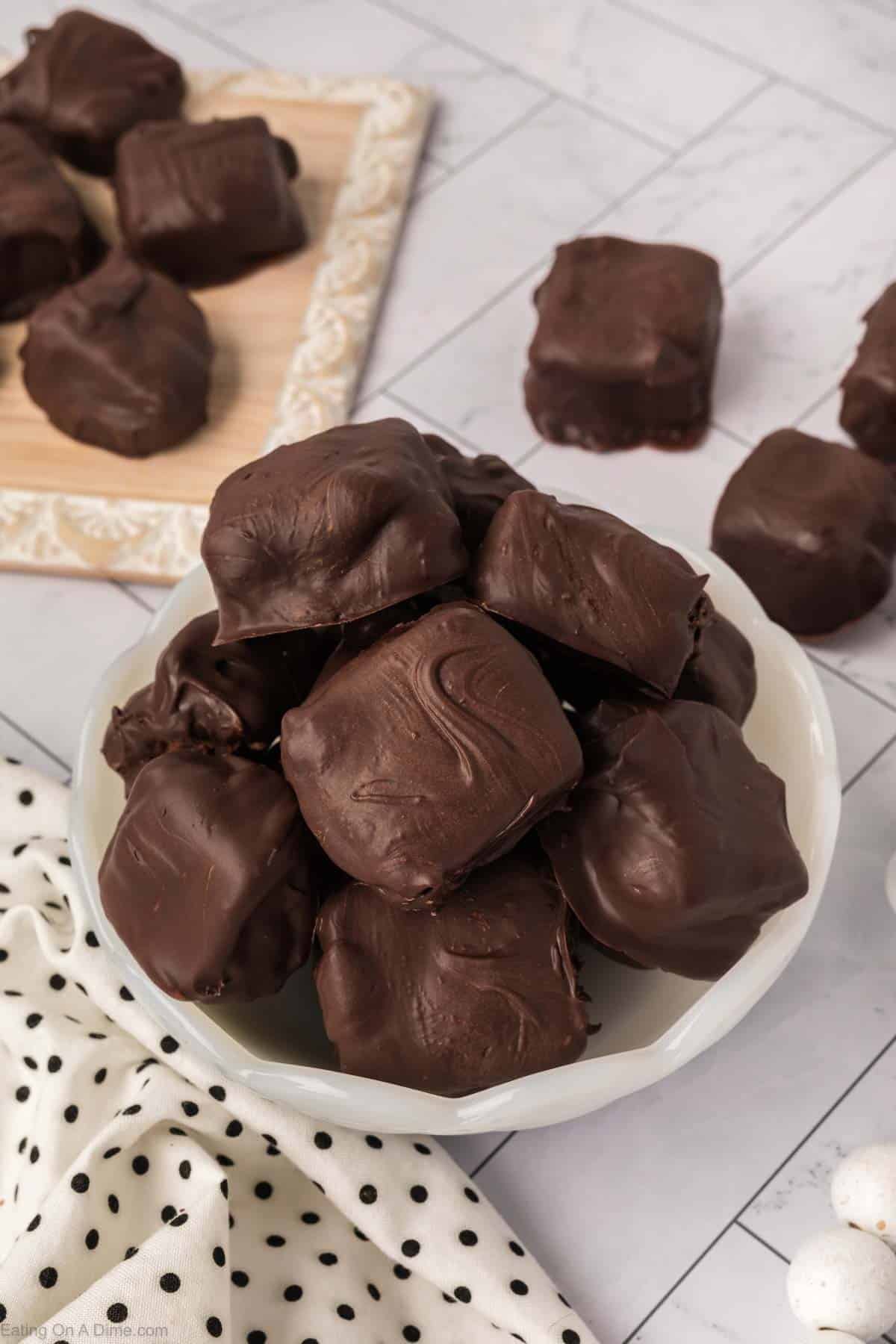 A white bowl filled with several chocolate-covered candy square treats sits on a polka dot cloth, with more treats on a wooden board and a tiled surface in the background.