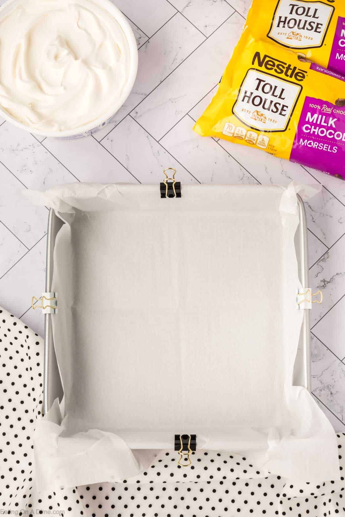 A square baking pan lined with parchment paper, clipped with binder clips, sits on a marble surface. Nearby are bags of Nestlรฉ Toll House chocolate morsels, a bowl of Cool Whip Candy mixture, and a polka dot cloth.