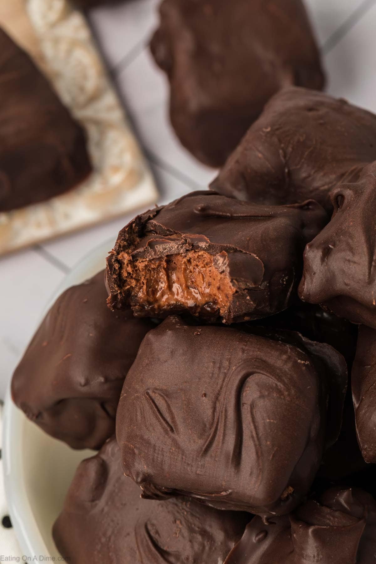 A close-up of chocolate-covered candy squares piled in a bowl, with one piece at the top showing a bite taken out, revealing a creamy Cool Whip and chocolate filling inside.
