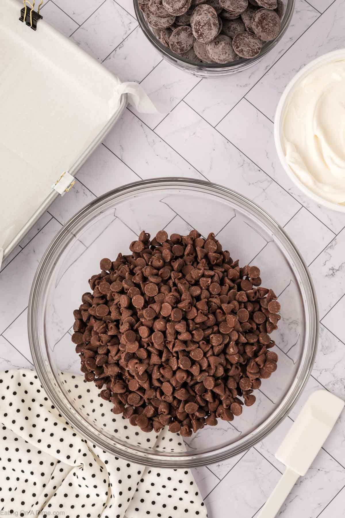 A glass bowl filled with chocolate chips sits on a tiled surface, surrounded by a spatula, a dotted cloth, a bowl of Cool Whip, and a bowl of candy pieces.